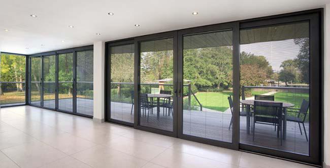 Spacious room with large sliding glass doors and expansive windows opening to a patio with outdoor table and chairs, overlooking a green lawn and trees.