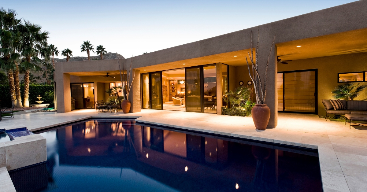 Modern house with large glass doors and expansive windows opening to a backyard patio and swimming pool, surrounded by palm trees and outdoor seating at sunset. Ideal for those seeking seamless room additions that maximize natural light and indoor-outdoor living.
