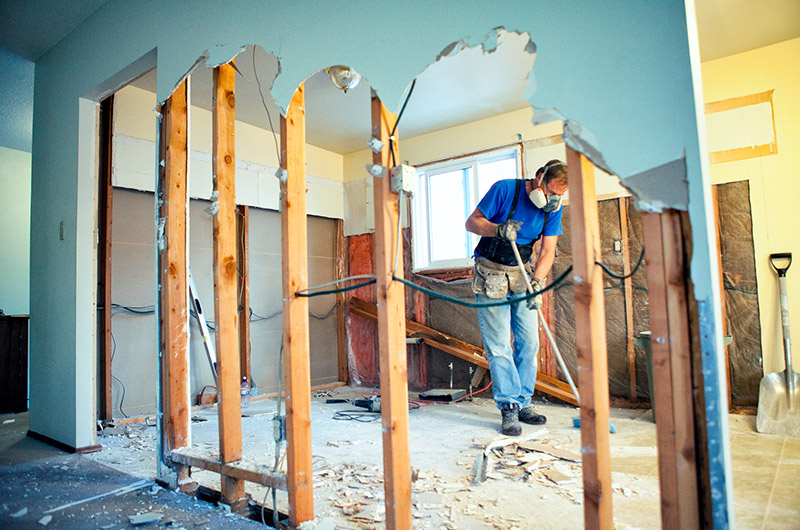 A person wearing a face mask works on demolishing an interior wall, with exposed wooden beams and debris on the floor—an essential step in revitalizing homes across Arizona through expert remodeling services.