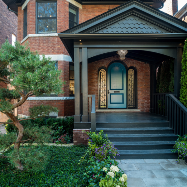A brick house in Arizona with a covered front porch, green exterior door replacement, black railings, and surrounding greenery including a small tree and shrubs.