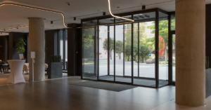 Modern lobby featuring large glass entrance doors and chic multi-slide doors, polished wooden floors, and stylish lighting. Tables and indoor plants add to the ambiance in the background.