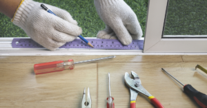 Gloved hands use a ruler to measure a window frame, amidst a screwdriver, pliers, and other tools on the wooden surface—ideal for those planning installations like multi-slide doors.