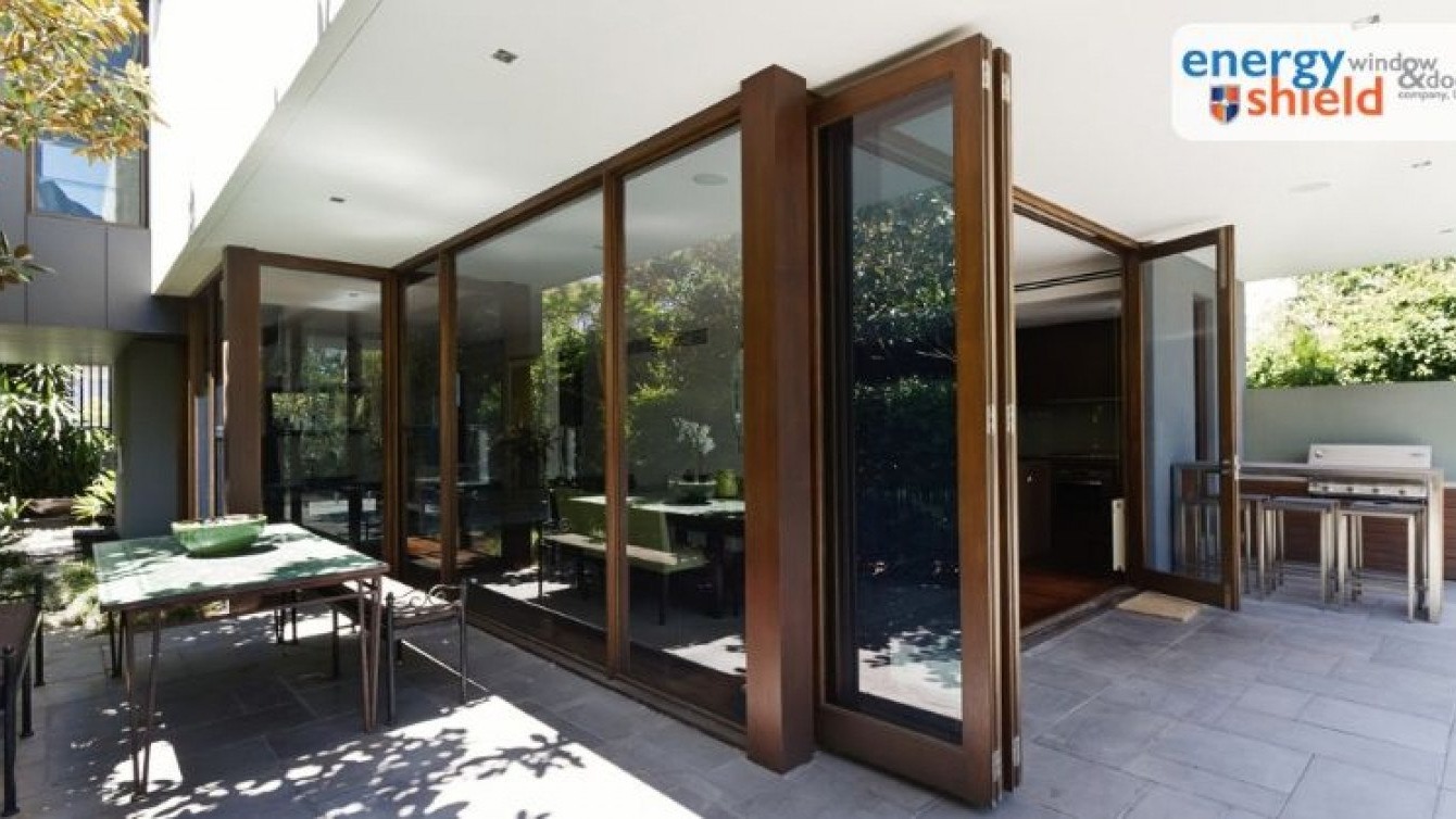 Large glass folding doors open from a modern interior dining area to an outdoor patio, seamlessly connecting indoor and outdoor living spaces in Arizona homes; the logo for Energy Shield Window & Door Company appears in the top right corner, highlighting their commitment to energy efficiency with beautifully crafted, energy-efficient doors.