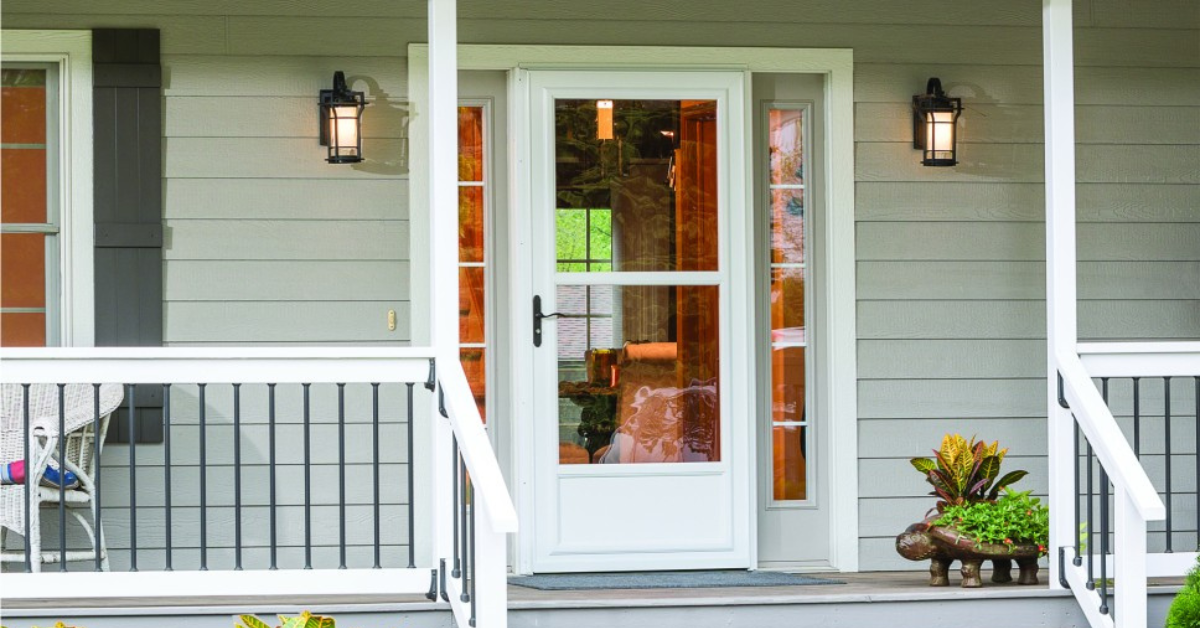 Front door of a Phoenix home featuring a white storm door, gray siding, two wall-mounted lantern lights, railing, and a planter with various plants on the porch.
