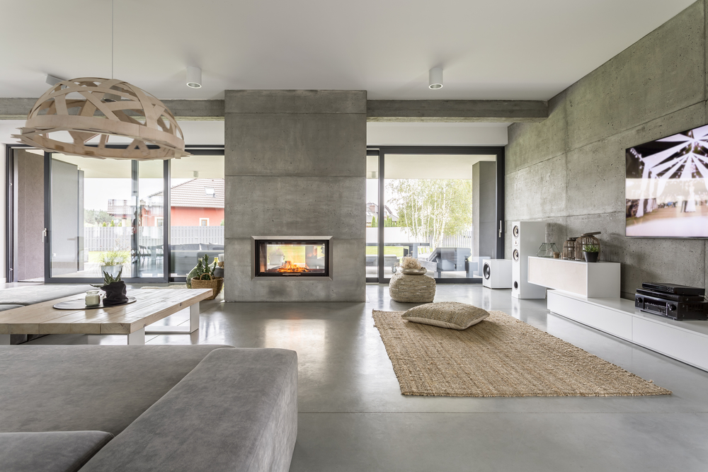Modern living room with concrete walls, a central fireplace, large windows that flood the space with natural light, a light gray sofa, a woven rug, low cushions, a wooden table, and a TV mounted on a white cabinet—perfect for seamless room additions or upgrading to stylish new doors and windows.